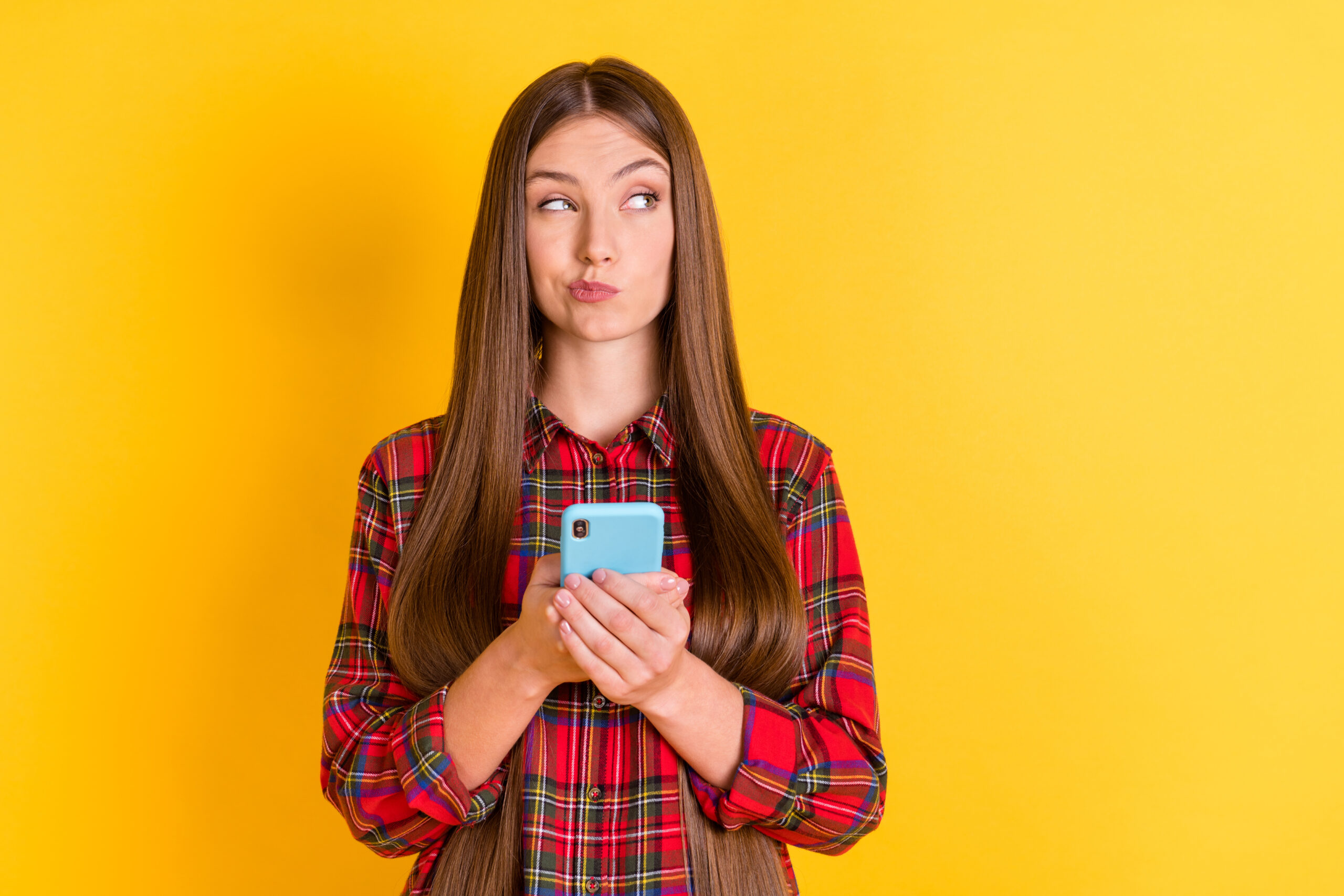 Portrait of attractive skeptic minded girl wearing checked shirt using device deciding copy space isolated over bright yellow color background