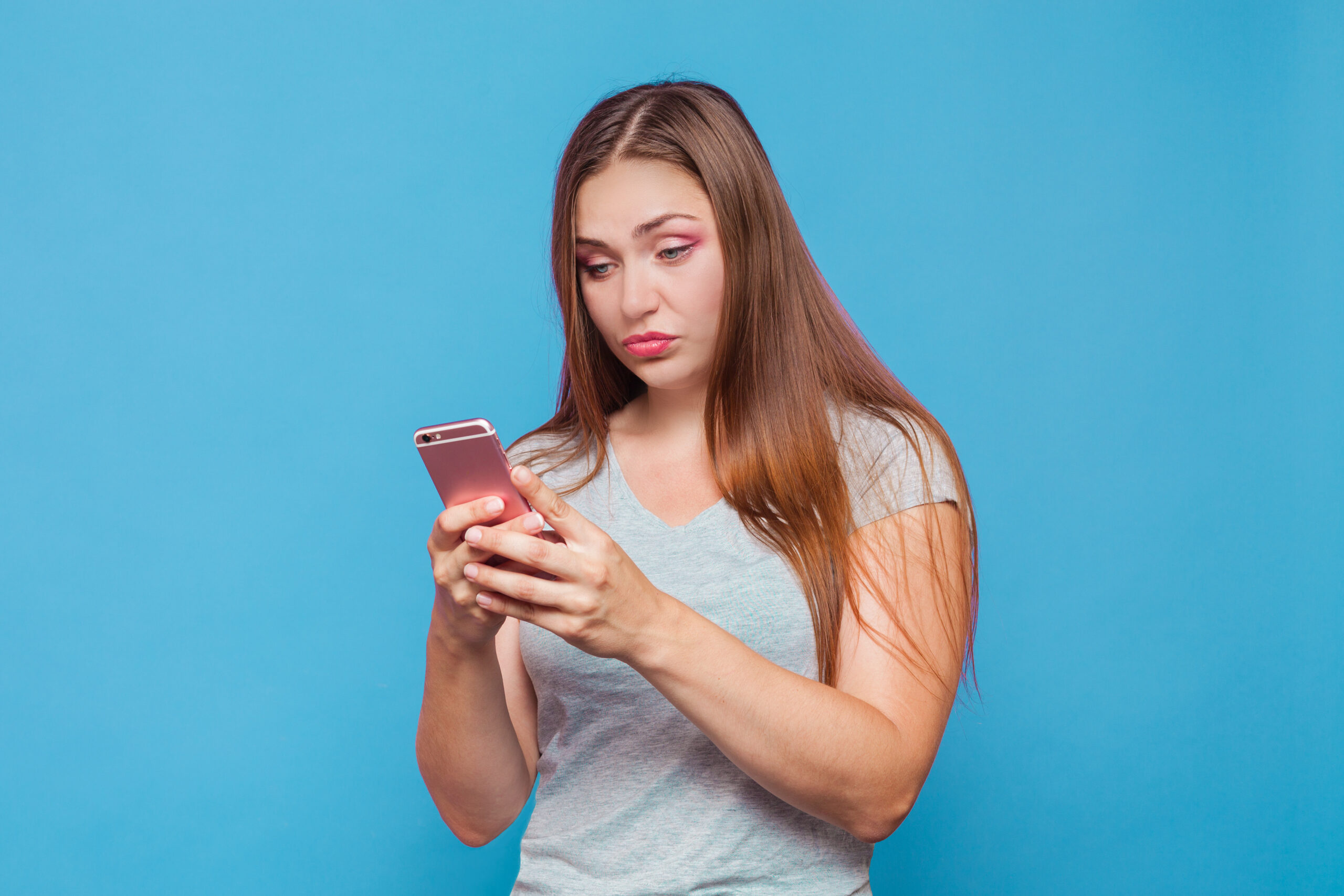 Young caucasian woman with brown hair and blue eyes looking to the pink phone with incredulous grimace, duckface. Blue background, studio, isolated