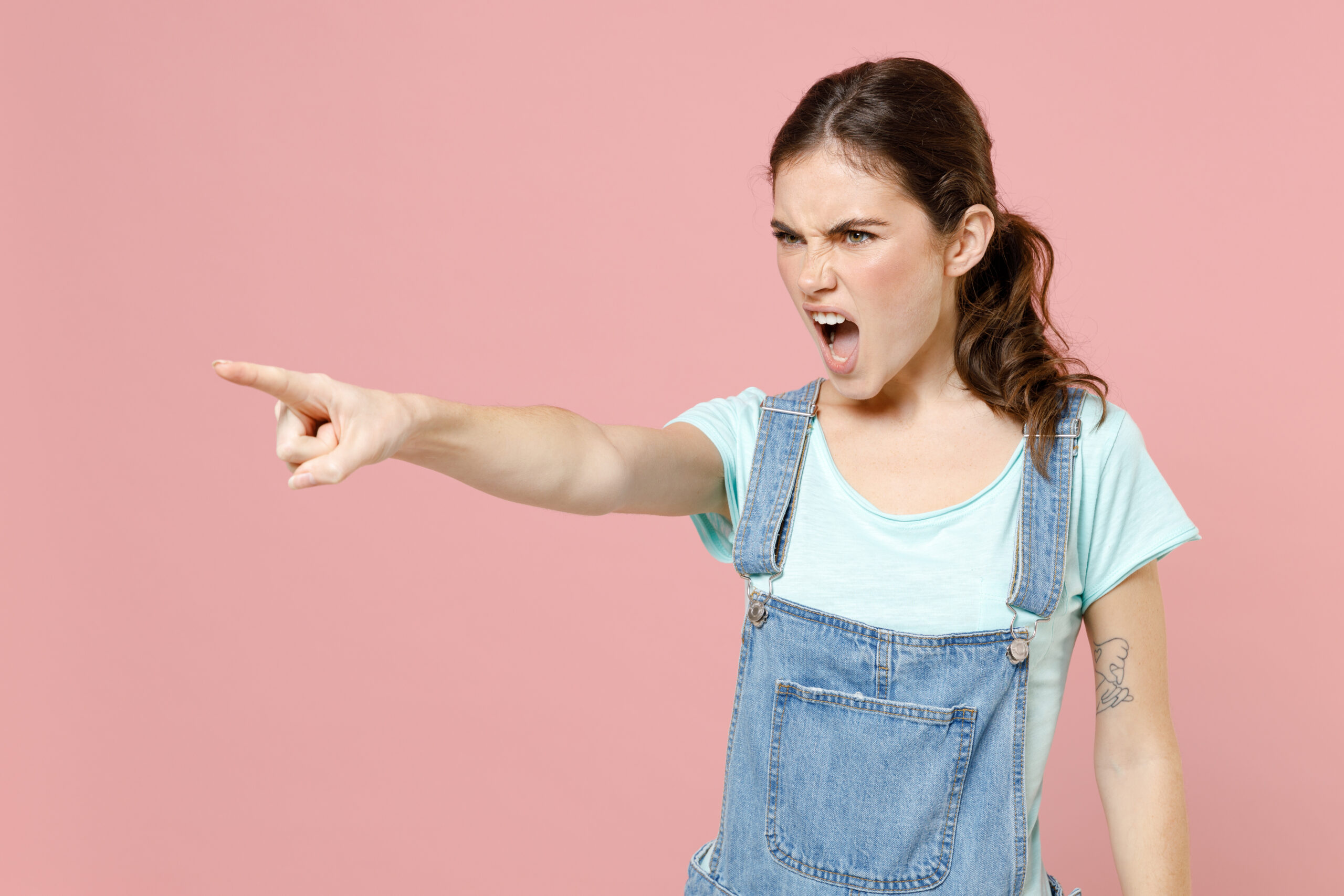 Young strict angry caucasian woman 20s wearing denim clothes blue t-shirt point index finger aside scream command do it isolated on pastel pink background studio portrait. People lifestyle concept