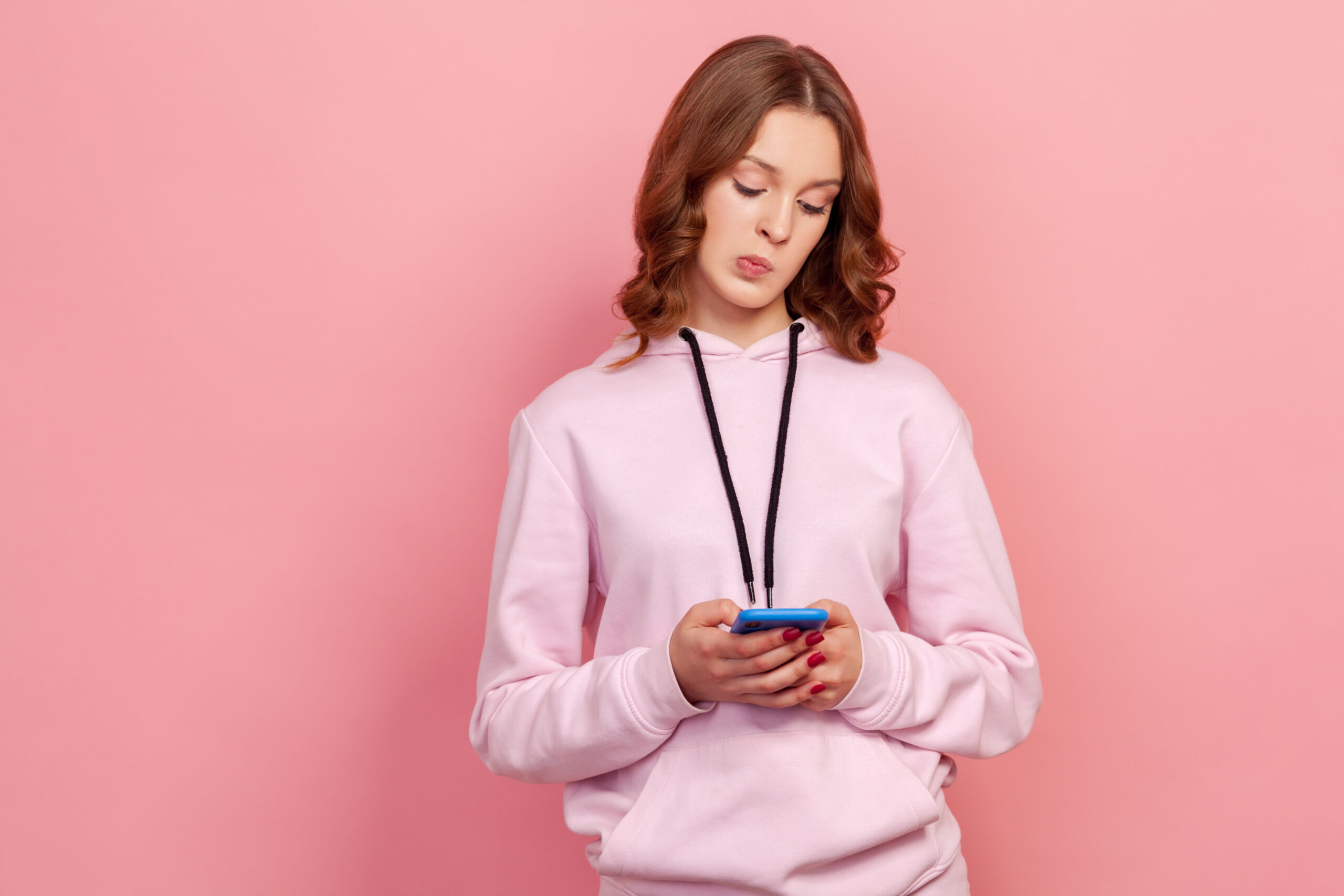 Portrait of pensive curly haired teenage girl in hoodie standing with mobile phone, thinking over message answer, having doubt suspicion feeling. Indoor studio shot isolated on pink background