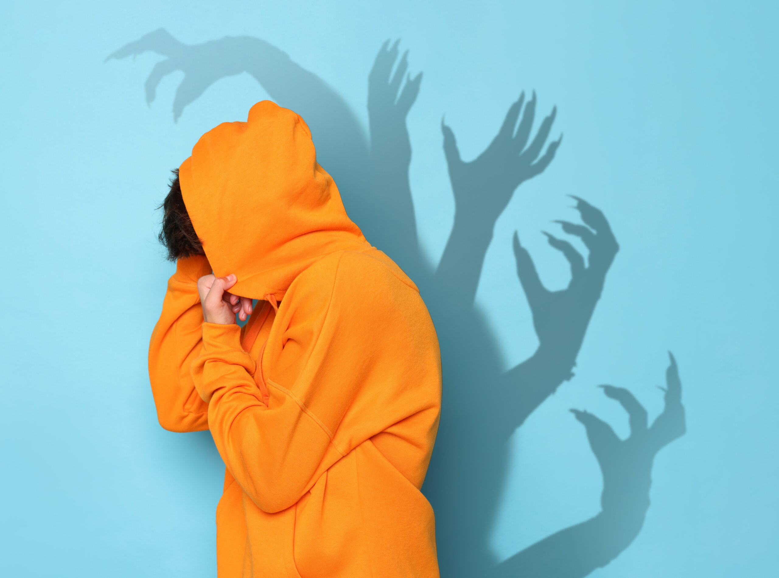 Scared teenage boy hiding on light blue background. Shadows of creepy hands behind him. Phobia, suffering from pathological fear