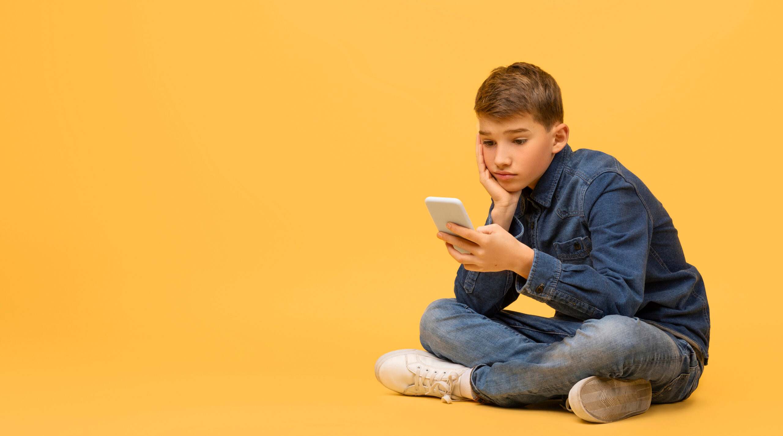 Bored teen boy using smartphone while sitting on floor in studio